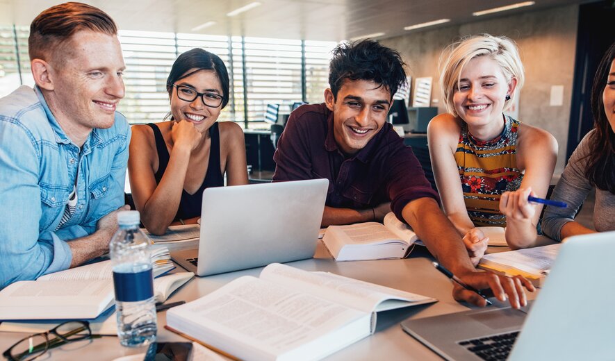 Weiterbildung für Auszubildende: Eine Gruppe Azubis sitzt in einer Lerngruppe vor einem Laptop. Ein junger Mann zeigt auf den Bildschirm von einem Laptop und erklärt den anderen Azubis etwas.